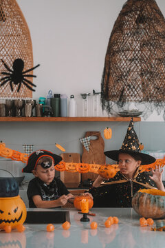 Portrait of a girl and her a little brother celebrating Halloween and decorating pumpkins in the kitchen. They look scary in witch and pirate costumes. Kids Halloween party.