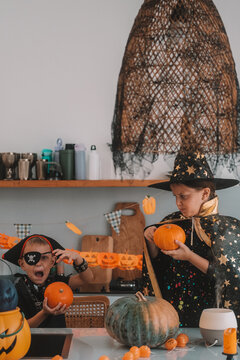 Portrait of a girl and her a little brother celebrating Halloween and decorating pumpkins in the kitchen. They look scary in witch and pirate costumes. Kids Halloween party.