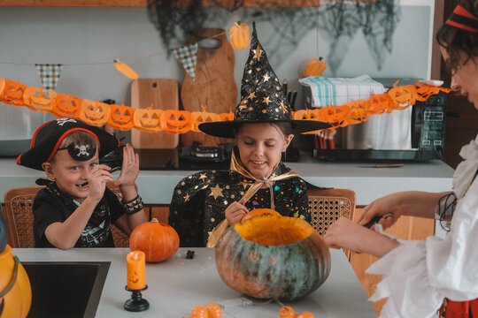 A portrait of a young girl and her younger brother celebrating Halloween and carving pumpkins in the kitchen with their mother's help. They are dressed in witch and pirate costumes. 
