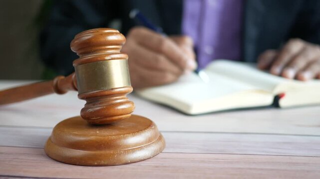 Judge's gavel on wooden table with person writing in notebook, concept of justice and law
