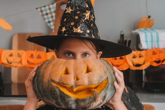 A portrait of a little girl dressed as a witch, holding a large pumpkin with a scary smile carved into it near her head. A children's Halloween party. The main autumn holiday.