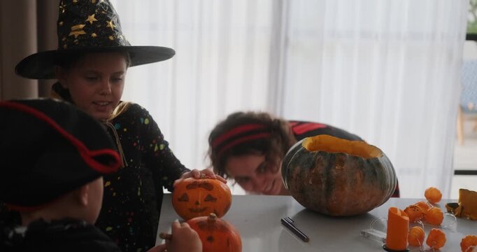 A portrait of a young girl and her younger brother celebrating Halloween and carving pumpkins in the kitchen with their mother's help. They are dressed in witch and pirate costumes. 
