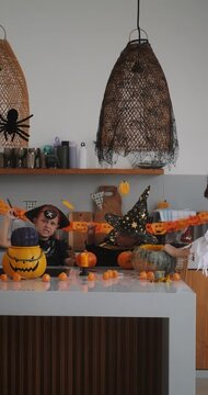 A portrait of a young girl and her younger brother celebrating Halloween and carving pumpkins in the kitchen with their mother's help. They are dressed in witch and pirate costumes. 