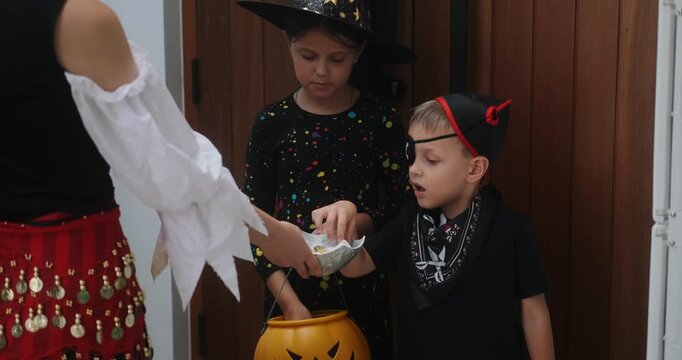 A portrait of a girl and her younger brother celebrating Halloween. They stand by the door with a pumpkin shaped basket and choose sweets. Trick or treat. Children's costume party.