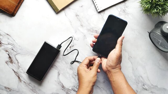 Top view of person's hands connecting a power bank to a black smartphone, charging on white marble table with office items