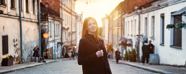 An adult woman stands confidently on a picturesque cobblestone street at twilight, surrounded by historic buildings and softly glowing street lamps. The atmosphere exudes charm and adventure. © Aleksandr