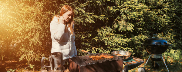 An adult woman stands at a barbecue grill in a backyard, focusing on the cooking process. Surrounded by lush greenery, she seems to enjoy the outdoor atmosphere and the act of grilling. © Aleksandr