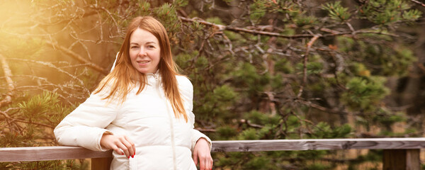 A woman stands relaxed by a wooden railing, surrounded by greenery in a tranquil outdoor environment. Her white coat contrasts beautifully with the natural background, highlighting a peaceful moment. © Aleksandr