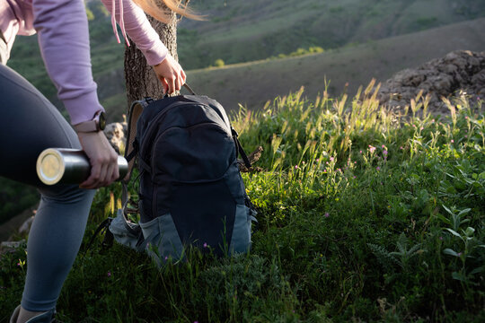 Woman traveler preparing to continue hiking by lifting her backpack in a scenic mountain meadow surrounded by wildflowers. Active lifestyle and adventure travel.