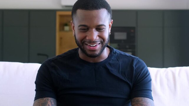 Overjoyed young african american man with tattoos watching sports on tv and celebrating a goal, showing pure excitement and happiness while sitting on a couch in his modern living room