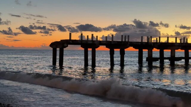 Old abandoned concrete pier on the shore with breaking ocean waves during sunset