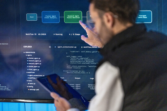 A product lead holding a tablet at a wall display keeps his palm open toward the live orchestration panel beside a bright blue wall panel during a late night shift.