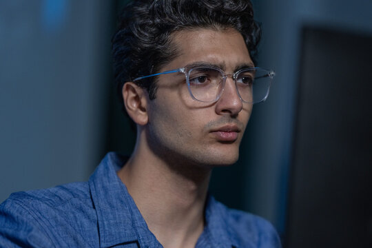 A young engineer in a blue shirt and clear glasses holds a steady look at the screen, the office dark behind in a tight portrait against the blue lit room, shot in low light.