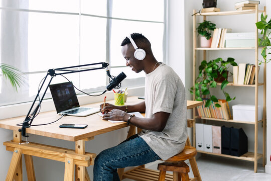 African man wearing headphones and using a microphone, recording podcast while writing notes at a desk indoors