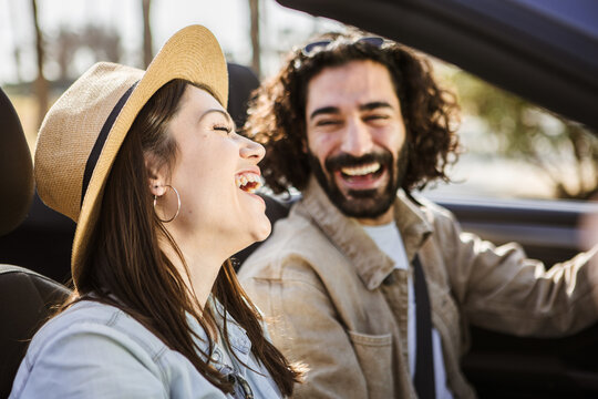 Young couple laughing while driving convertible car on sunny day. Man and woman enjoying carefree road trip during summer vacation. Travel and lifestyle concept.