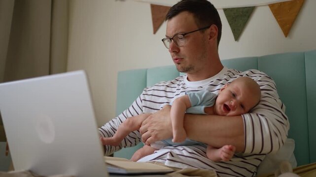 Young busy father on paternity leave trying to work as a freelancer from a home office, holding a crying newborn baby and feeling stressed with the challenges of multitasking