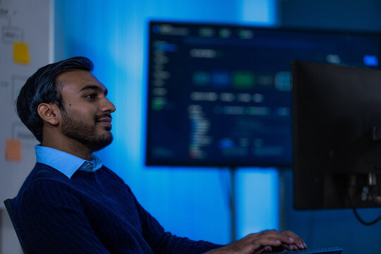 A male technology consultant in a tailored dark jacket and open collar keeps his eyes on the monitor as new alerts arrive while critical systems run around the clock