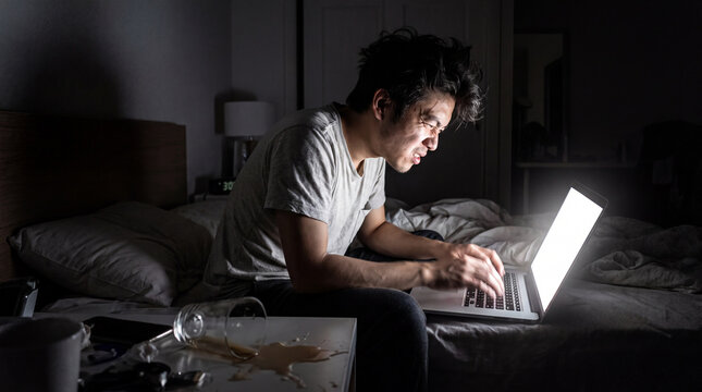 exhausted DevOps engineer abruptly woken up for an on-call emergency. A young Asian man is sitting up in a messy bed at 3 AM in a completely dark room.