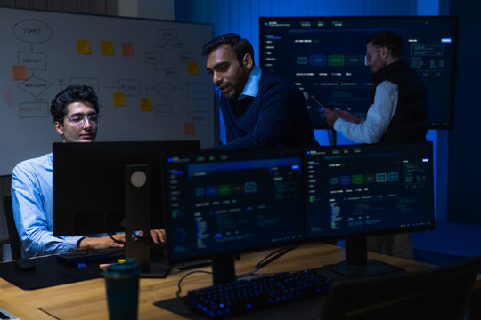 A junior male software engineer in a pale blue shirt holding a digital tablet lean in together as the metrics finally return to normal while critical systems run around the clock