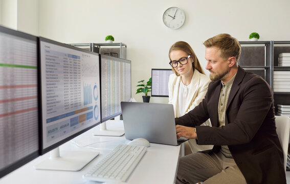 Analysts and businesswoman review finance data on monitors. Colleagues work at a laptop in bright office comparing spreadsheets and charts to assess analytics. Teamwork fuels data led choices.