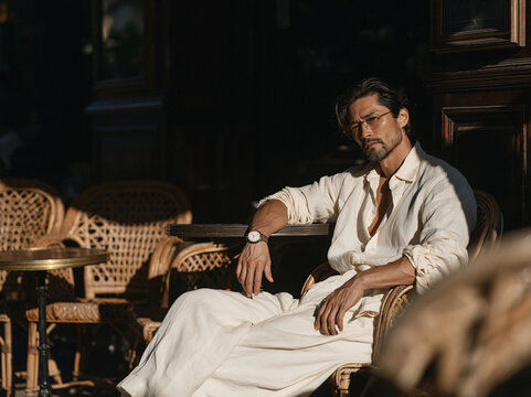 Stylish man in white linen suit and sunglasses sits at vintage Parisian caf&eacute; bathed in warm golden afternoon side light.