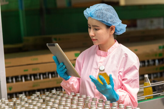 Asian female scientist working on tablet computer check quality of drinking beverage on conveyer belt before distribution to market.