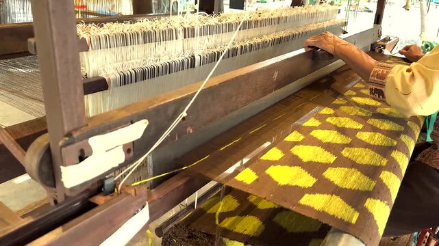 Artisanal hand weaver working on traditional wooden loom to create intricate yellow and brown textile patterns.