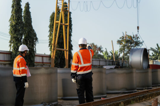 Engineers control crane lifting prefabricated steel cylinder at outdoor factory yard. Concept of heavy manufacturing, infrastructure development, industrial logistics and engineering management.