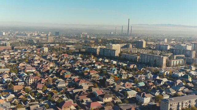 Aerial panorama of Bishkek, Kyrgyzstan, showing residential neighborhoods beside factories and a tall chimney within the urban landscape.
