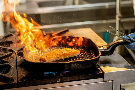 Seared salmon fish cooks in flaming cast-iron pan. Black cast-iron pan sits on gas stove. Bright flames surround sizzling salmon fish inside the pan. Blue-gloved hand holds tongs, flipping the fish.