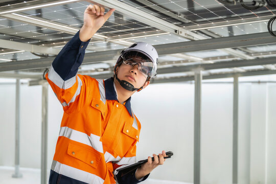 Young engineer in orange uniform and white helmet examines solar panels and metal beam structure reviewing installation details on tablet. expression shows concentration of new gen engineer