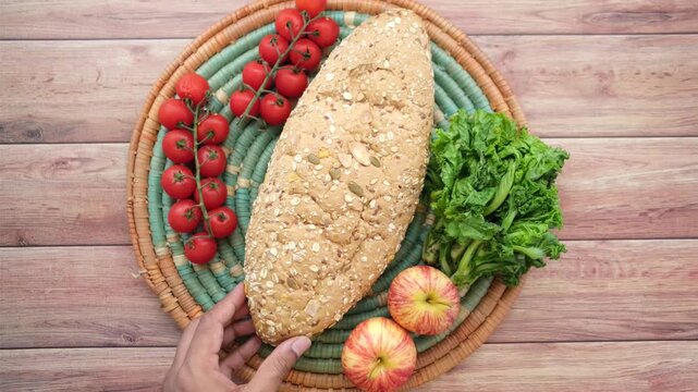 A person placing homemade multigrain bread loaf with fresh vegetables and fruits on a wooden table, top view dynamic footage