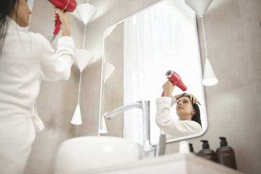 A woman wearing a bathrobe uses a hairdryer while looking in the mirror. The chic bathroom features elegant lighting, minimalistic design, and various hair care products on display