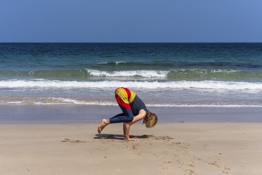 A woman doing yoga on a sandy beach by the sea. Kakasana or crow. Treen Cove, Cornwall, England, Great Britain