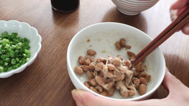 朝の食卓で白いお皿に入った納豆をお箸で混ぜているシーンのアップ