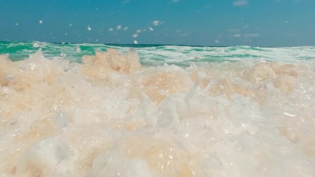 Slow motion low angle shot of turquoise ocean wave breaking on tropical golden sand beach under bright blue sky.
