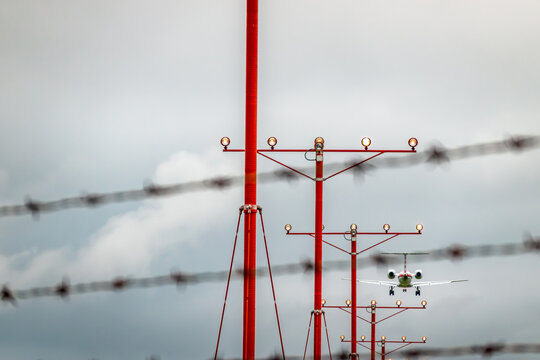 Commercial aircraft on final approach framed by security barbed wire fencing and bright red airport approach lighting systems