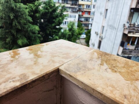 Wet balcony ledge after rain with city apartment buildings and trees in background, stone parapet closeup in autumn weather, urban rooftop view, autumn concept