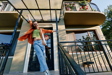 Young woman rejoices outside her apartment building, raising arms happily. She wears casual orange...