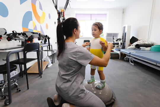 Cute boy with cerebral palsy works on balance and coordination with physical therapist in a clinic room standing in balance hemisphere