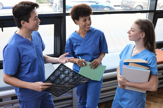 Multiracial medical students in blue scrubs discussing a brain MRI scan near a window in a library. Neurosurgery diagnostics, stroke screening, and clinical neurology study