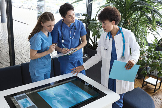 Senior physician demonstrating pelvic bone radiography on a smart touch console to attentive medical residents in a hospital lobby