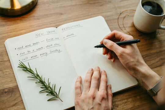 New Year resolutions list in open notebook, person writing goals on wooden desk, cozy lifestyle planning with coffee and rosemary