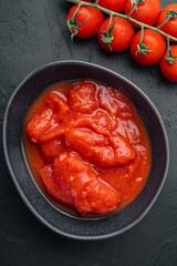 Obraz premium Canned tomato chunks in dark ceramic bowl beside fresh vine tomatoes on black textured surface under soft directional lighting