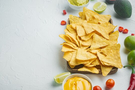 nachos chips piled high on wooden serving board with creamy cheese dip guacamole avocado slices lime wedges cherry tomatoes red chili peppers and whole green limes arranged on textured white