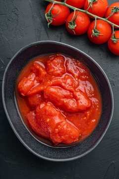 Canned tomato chunks in dark ceramic bowl beside fresh vine tomatoes on black textured surface under soft directional lighting