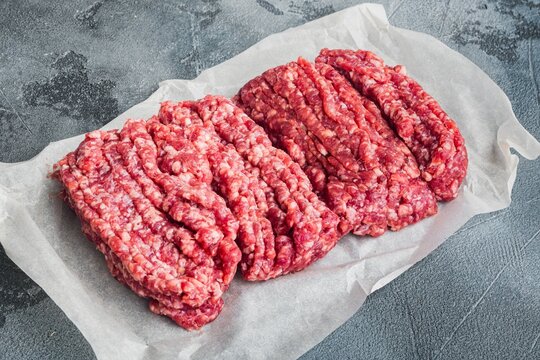 Two portions of raw minced beef with visible fat marbling arranged on crumpled parchment paper against a textured gray surface under even studio lighting