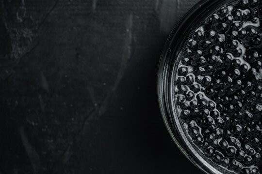 Black caviar in a glass jar with glossy surface and textured dark background, top view flat lay with ample copy space for text, close-up still life composition highlighting the round glistening beads