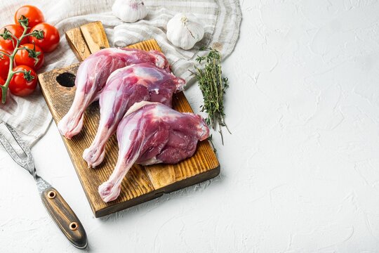 Three raw duck legs resting on a wooden cutting board beside fresh garlic cloves thyme sprigs and vine tomatoes all arranged on a textured white stone surface under soft even lighting with ample