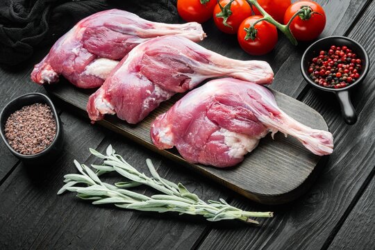 Three raw duck leg meat thighs resting on a dark wooden cutting board beside fresh red tomatoes still on vine, small bowls of mixed peppercorns and coarse grain seasoning